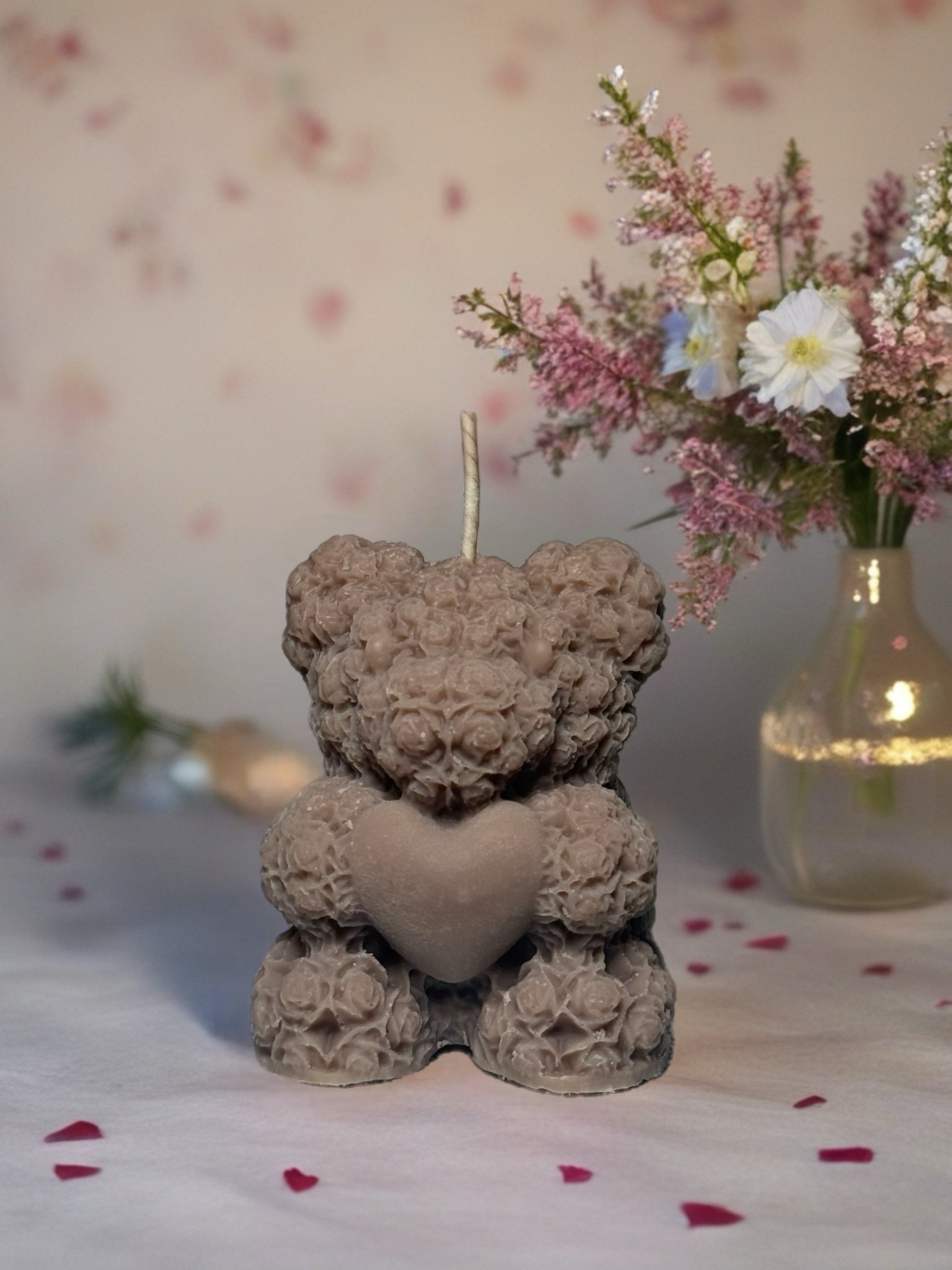 Decorative candle shaped like a teddy bear with a heart, placed on a surface with flowers in the background.